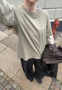 Khaki overhemd met lange mouwen van lichte stof, gecombineerd met donkerdere wijde broekspijpen. Zwarte tas in één hand, met kasseien op de achtergrond.