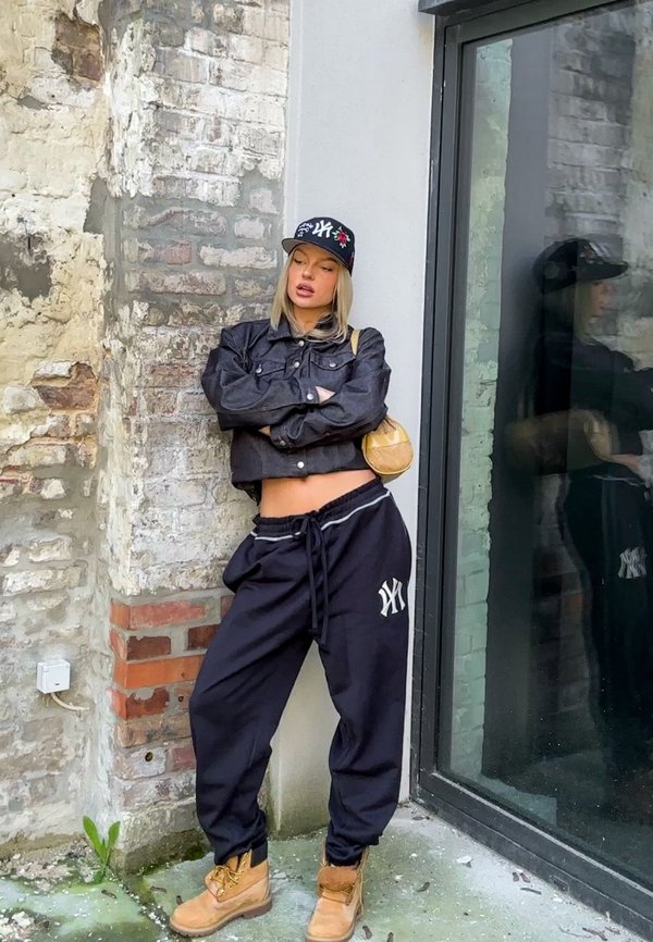 Black cropped denim jacket, black drawstring sweatpants with a white logo, tan boots, and a black cap with floral accents. Urban backdrop.