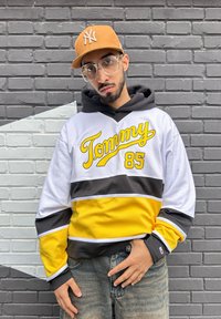 Felpa a rete bianca e gialla con logo "Tommy 85", strisce nere e gialle, abbinata a jeans azzurri chiari e un cappellino da baseball color sabbia.