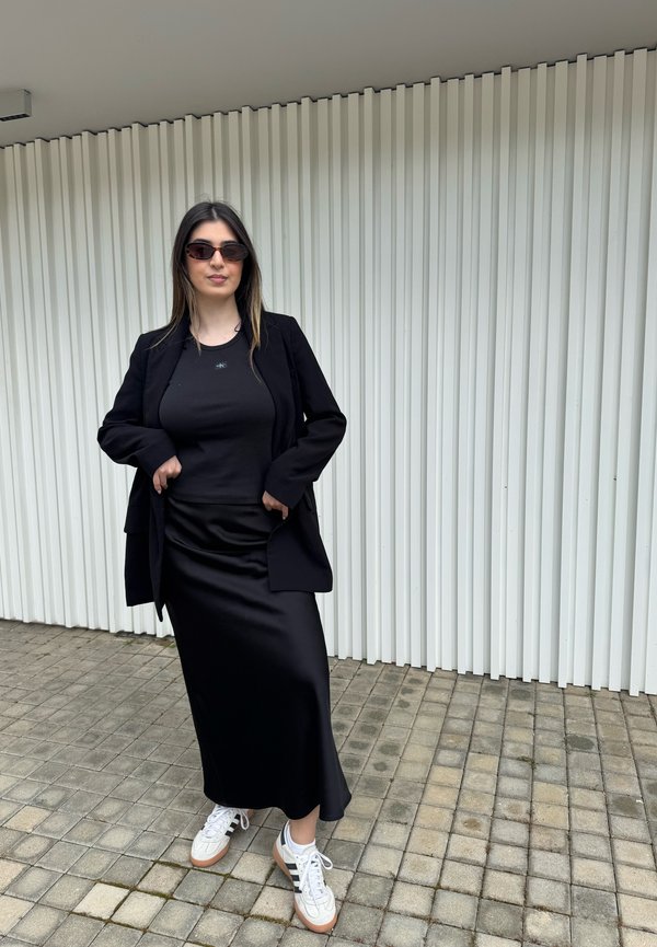 Junge Frau in schwarzem Blazer, schwarzem Oberteil, langem schwarzem Rock, weißen Sneakers und Sonnenbrille steht auf gepflastertem Gehweg vor einer weißen vertikalen Paneelwand.