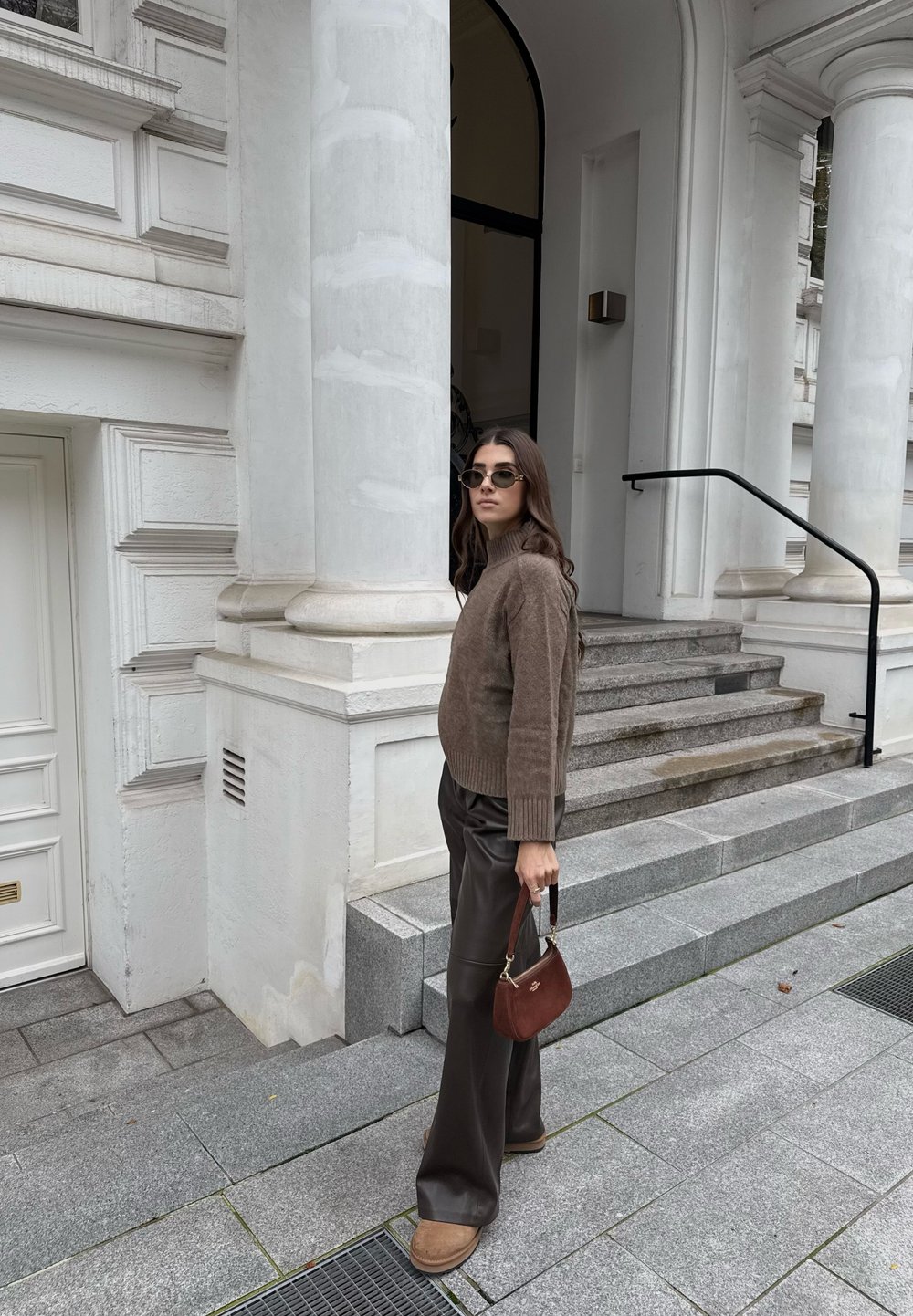 Rjav pleten pulover, temno usnjeni široki hlače, majhna rjava torbica in sončna očala, ki stoji ob kamnitih stopnicah in belih kolumnah.