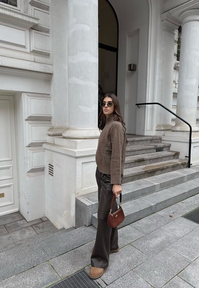 Suéter de punto marrón, pantalones de cuero oscuros de pierna ancha, un pequeño bolso marrón y gafas de sol, de pie junto a una escalera de piedra y columnas blancas.