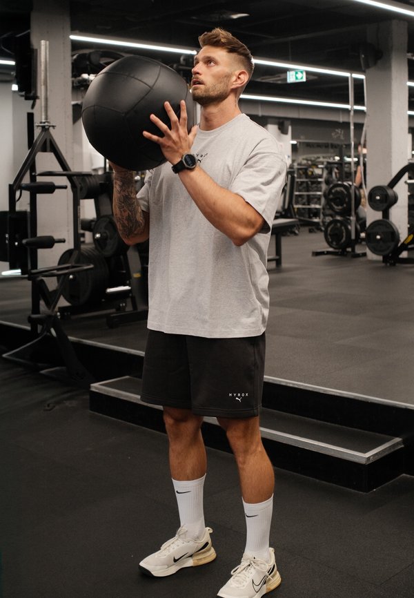 Homme portant une chemise grise et un short noir tenant un ballon médicinal noir, se préparant à faire de l'exercice dans une salle de sport avec des poids en arrière-plan.