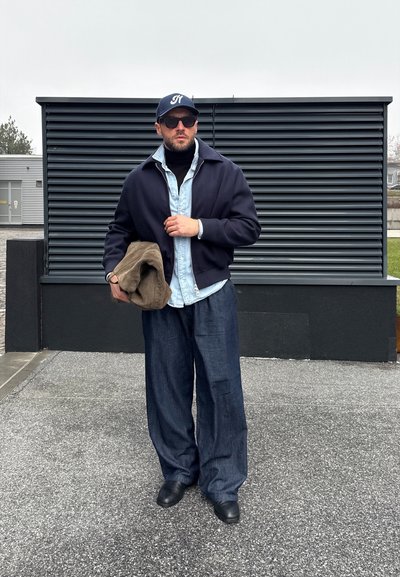 Hombre con gafas de sol oscuras, chaqueta azul marino, camisa de mezclilla clara, pantalones anchos oscuros y gorra, sosteniendo una prenda marrón doblada al aire libre.