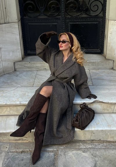 Mujer sentada en escalones de mármol vistiendo un abrigo largo marrón, botas hasta la rodilla, gafas de sol, guantes y una diadema, junto a un bolso marrón.