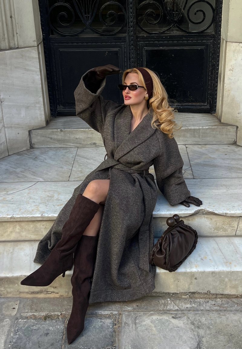 Femme assise sur des marches en marbre portant un long manteau marron, des bottes hautes jusqu'aux genoux, des lunettes de soleil, des gants et un bandeau, à côté d'un sac à main marron.