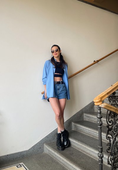 Camisa azul clara de gran tamaño, top negro corto, pantalones cortos de mezclilla, botas negras con cordones. Escalera de piedra con barandilla de madera y suelo con estampado.