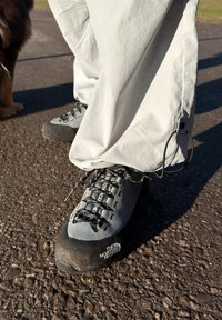 Zapatos de senderismo grises con detalles negros y suelas resistentes, con un sistema de cordones y logotipo. Pantalones blancos y sueltos cubren parcialmente los zapatos.