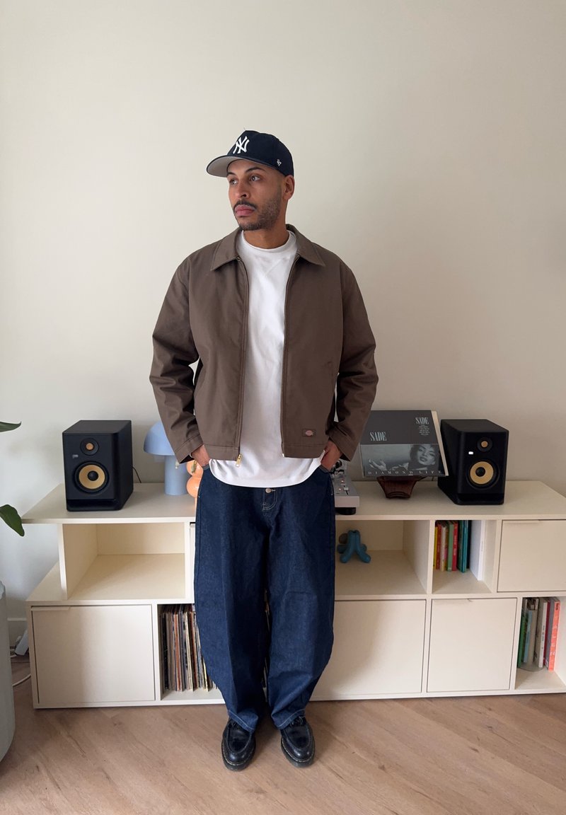 Homme portant une veste marron, une chemise blanche, un jean ample et une casquette noire, debout devant une étagère blanche avec des enceintes et des disques vinyles.