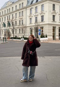 Καφέ ψεύτικο παλτό, κόκκινος λαιμόκοψος, τζιν παντελόνι, μαύρη τσάντα με κόκκινη λεπτομέρεια, μαύρα και άσπρα αθλητικά παπούτσια. Αστική τοποθεσία με ιστορικό κτίριο.