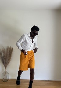 Man wearing white button-up shirt, bright orange shorts, black socks, and shoes holding a small bottle, standing beside a vase with dried pampas grass.