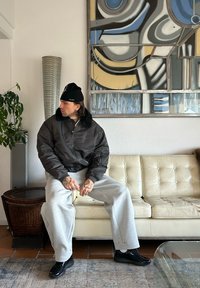 Veste bomber rembourrée noire avec col en fourrure, pantalon de survêtement gris, et baskets noires brillantes, assis sur un canapé en cuir capitonné couleur crème.