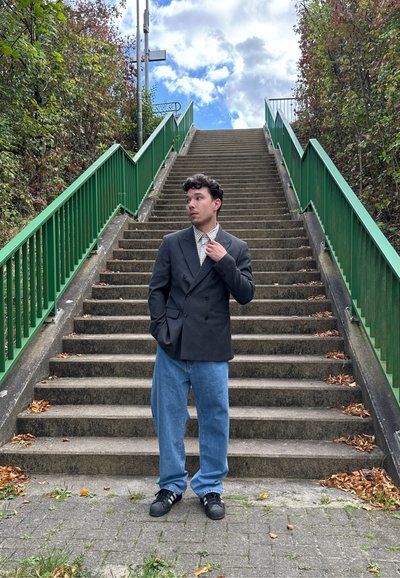 Blazer negro de doble botonadura sobre una camisa de cuadros, combinado con unos vaqueros de pierna ancha de color azul claro. De pie en unos escalones de piedra, con barandillas verdes visibles.