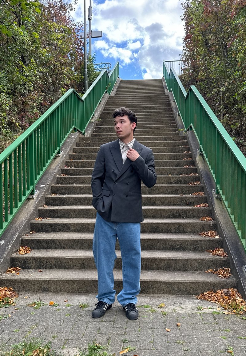 Blazer preto de dois botões sobre uma camisa de xadrez, combinado com jeans de perna larga azul claro. Em pé sobre degraus de pedra, com corrimãos verdes visíveis.