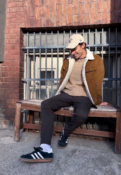Chaqueta de pana marrón con cuello de borreguito blanco, suéter de punto beige, pantalones negros y zapatillas de ante verdes con rayas blancas.