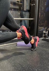 Svart og rosa treningssko med teksturert overflate, rund form og polstrede såler, plassert på et treningsgulv med treningsutstyr.