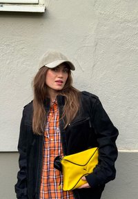 Crna jakna s prednjim patentnim zatvaračem i džepovima, narančasta i bijela košulja na kockice, siva kapa i žuta PUMA torbica s crnim detaljem.