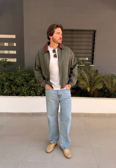 Hombre con atuendo casual que lleva chaqueta verde, camisa blanca, jeans azul claro, zapatos color beige y gafas de sol colgando del cuello, de pie junto a plantas.