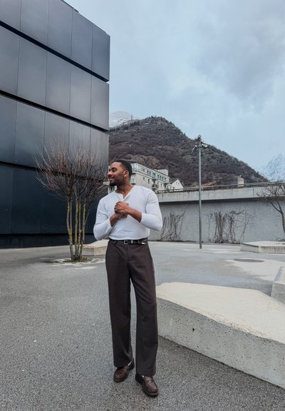 Hombre con camisa blanca de manga larga y pantalones oscuros de pie al aire libre cerca de un edificio negro, árboles sin hojas, estructuras de hormigón y montañas al fondo.
