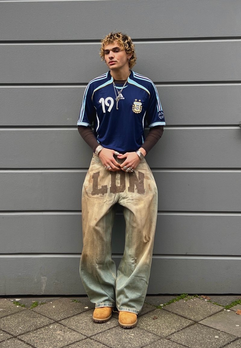 Jeune personne aux cheveux bouclés se tenant contre un mur gris, portant un maillot de sport bleu, un pantalon ample avec le texte « LION », et des bottes couleur sable.