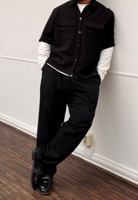 Chemise noire à manches courtes boutonnée avec deux poches poitrine, portée sur une chemise blanche à manches longues, associée à un pantalon noir et des chaussures noires brillantes.