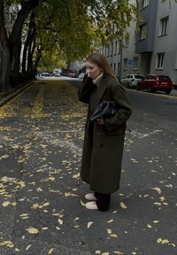 Μακρύ ελιάς παλτό πάνω από κρέμα ζιβάγκο, συνδυασμένο με μαύρη τσάντα και ανοιχτόχρωμα παντοφλάκια, σε δρόμο καλυμμένο με φύλλα.