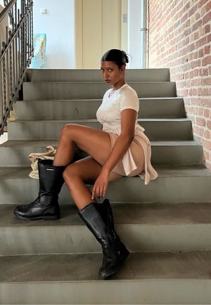 Botas negras hasta la rodilla, pantalones cortos beige claro y una camiseta blanca con detalles de logo, sentado en unas escaleras grises con un fondo de pared de ladrillo.