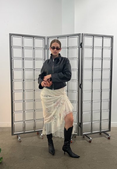 Chaqueta corta de cuero negro, falda crema con estampado floral y botas negras hasta la rodilla. El fondo presenta una pantalla de bloques de vidrio plegables.