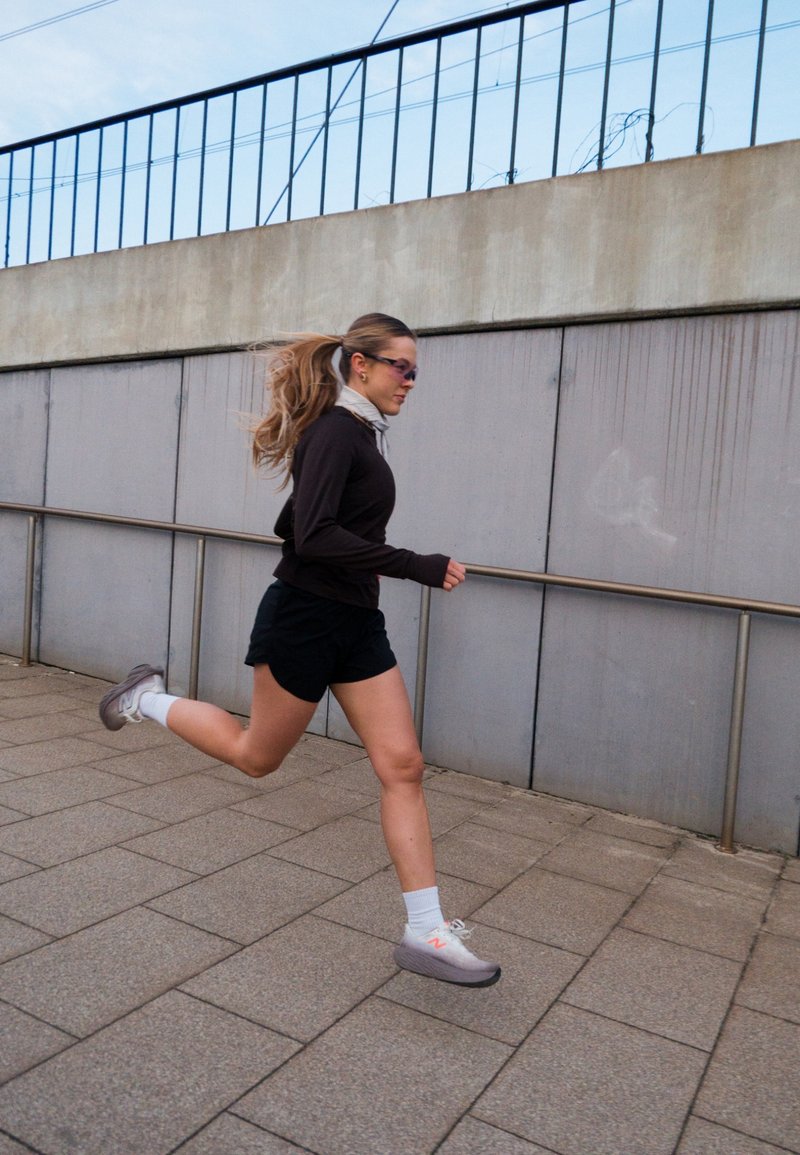 Femme vêtue de vêtements de sport noirs courant sur un chemin urbain pavé à côté d'un mur en béton avec une rambarde métallique.
