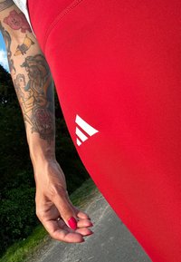 Červené sportovní legíny vyrobené z elastického materiálu, s bílým logem Adidas na spodní straně. Hladká textura a přiléhavý střih.