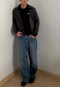 Νέος άνδρας που φοράει μαύρο δερμάτινο μπουφάν, μαύρο πουκάμισο, φαρδιά μπλε τζιν και μαύρα παπούτσια, ακουμπώντας σε λευκό τοίχο.