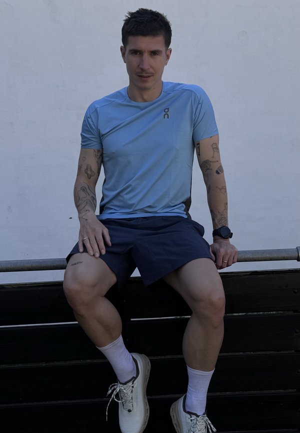 Homme en chemise bleu clair et short foncé avec des tatouages, assis sur un banc contre un mur uni, portant des baskets blanches et une montre connectée.