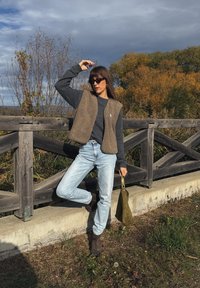 Colete de lã castanho sobre um suéter cinza, jeans azul claro e botas de tornozelo castanhas. Bolsa verde e corrimão de madeira em um ambiente natural.