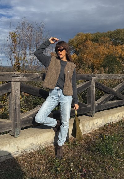 Chaleco de forro polar marrón sobre un suéter gris, jeans azul claro y botas de tobillo marrones. Bolso verde y barandilla de madera en un entorno natural.