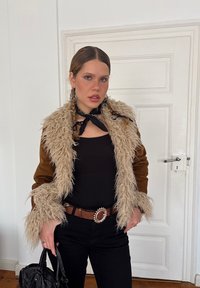 Giovane donna con capelli intrecciati che indossa una giacca in shearling marrone e beige, top nero, pantaloni neri, cintura marrone e tiene una borsa nera in un ambiente interno.