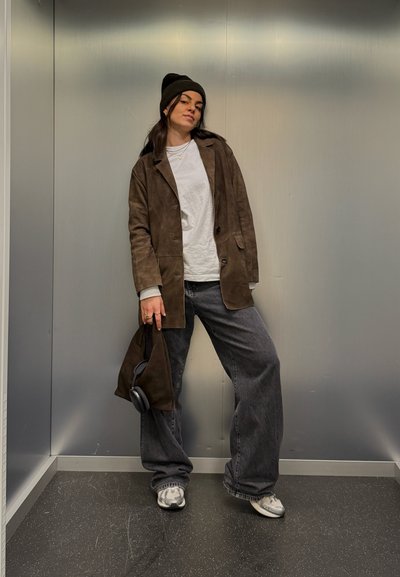 Blazer de ante marrón sobre una camisa blanca, combinado con pantalones anchos de mezclilla gris oscuro y zapatillas plateadas. Los accesorios incluyen un bolso oscuro y unos auriculares.