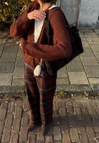 Bruine gebreide cardigan, wit topje, geruite broek in tinten bruin, zwarte leren handtas, grijze sleutelhanger met pompon-detail, bakstenen straat.