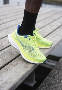 Lys gul joggesko med mesh-ovdel, blå detaljer og hvit såle. Brukt med en svart sokk, plassert på en trebrygge.