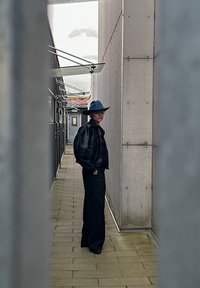 Μαύρο δερμάτινο μπουφάν πάνω από μαύρο τοπ, φαρδιά μαύρα παντελόνια και καπέλο με φαρδύ γείσο σε μπλε χρώμα; στέκεται σε έναν στενό, τσιμεντένιο διάδρομο.