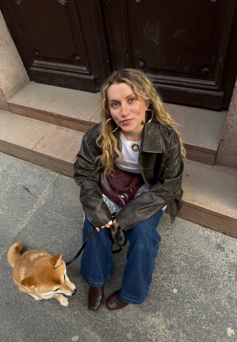 Jeune femme aux cheveux blonds ondulés assise sur des marches en pierre tenant un petit chien en laisse, portant une veste en cuir et de grandes boucles d'oreilles créoles.