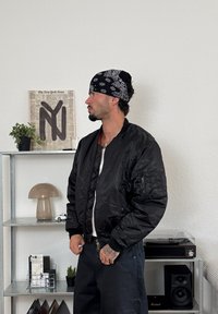 Zwarte bomberjack met een ritssluiting aan de voorkant en geribde boorden, gedragen over een wit shirt. De achtergrond bevat een glazen plank met decoratieve items.