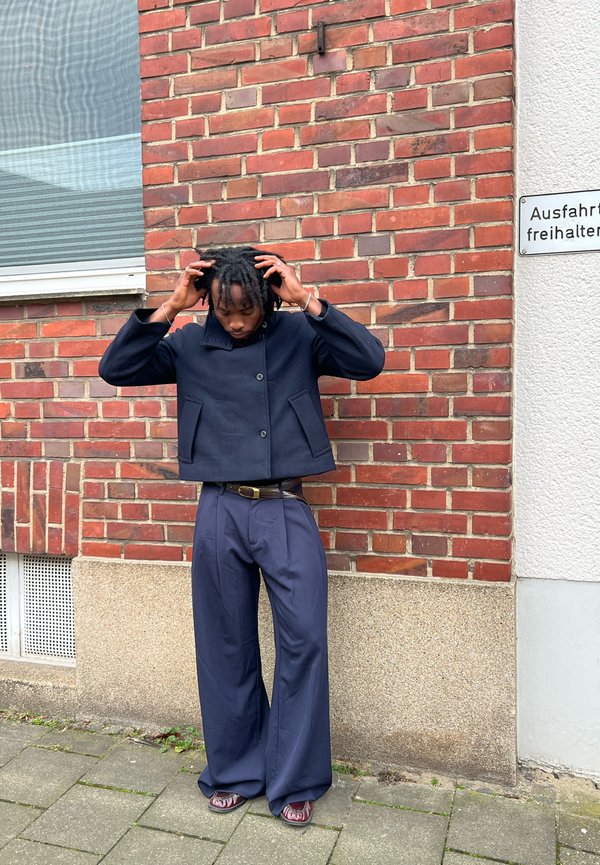 Merisotilasleikkaus takki suurilla etutaskuilla, yhdistettynä leveälahkeisiin merisotilashousuihin. Ulkoilma-asetus teksturoitu tiiliseinä taustalla.