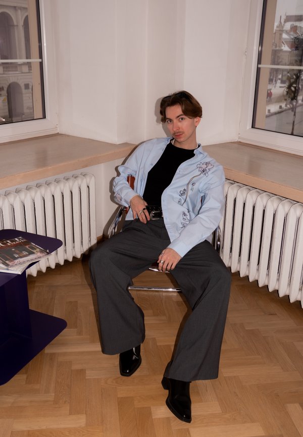 Mladá osoba nosící černé boty, šedé široké kalhoty a světle modrou vyšívanou košili, sedící v rohové místnosti s dřevěnou podlahou a radiátory.