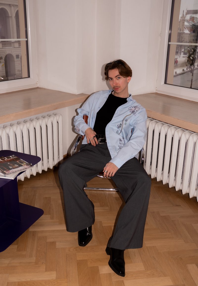 Jeune personne portant des bottes noires, un pantalon large gris et une chemise brodée bleu clair, assise dans une pièce d'angle avec un sol en bois et des radiateurs.