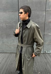 Vīrietis, kura mugurā ir pelēks trencs un melns turtlenecks, saulesbrilles un kas tur siksnu pāri plecam, stāv pelēka flīžu sienas priekšā.