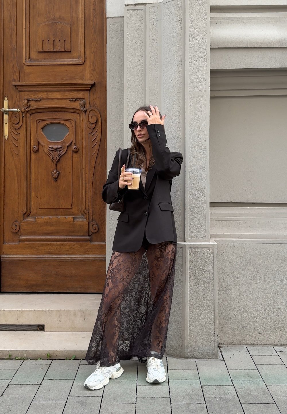 Čierny blazer cez priesvitnú čipkovanú sukňu, biele tenisky, držiac ľadovú kávu. Stojí pred drevenými dverami s dekoratívnymi detailmi.