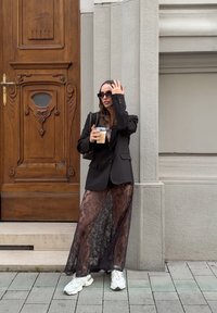 Čierny blazer cez priesvitnú čipkovanú sukňu, biele tenisky, držiac ľadovú kávu. Stojí pred drevenými dverami s dekoratívnymi detailmi.