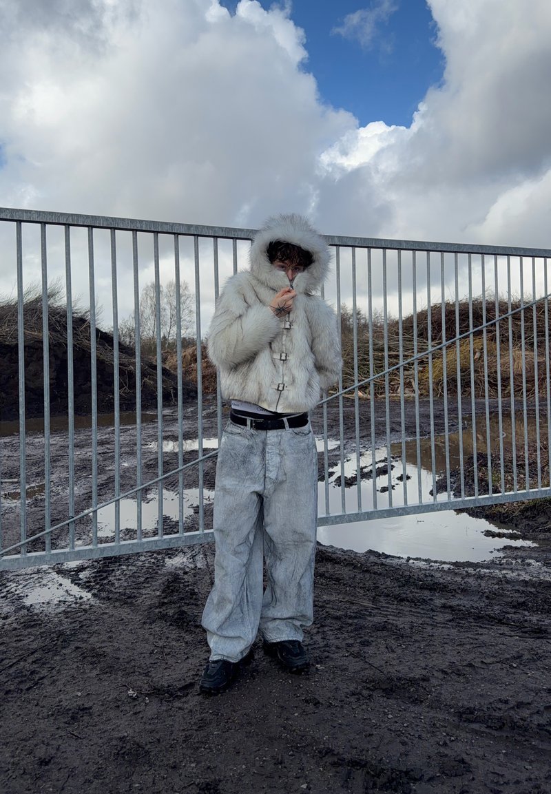 Άτομο που φοράει λευκό γούνινο μπουφάν με κουκούλα και φαρδιά ανοιχτογρίζα παντελόνια στέκεται σε λασπωμένο έδαφος κοντά σε μεταλλική πύλη κάτω από συννεφιασμένο ουρανό.