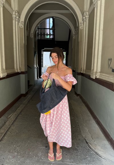 Vestido rosa con lunares, diseño sin mangas, sosteniendo un bolso oscuro con un acento amarillo y artículos verdes dentro, situado en un pasillo arqueado.