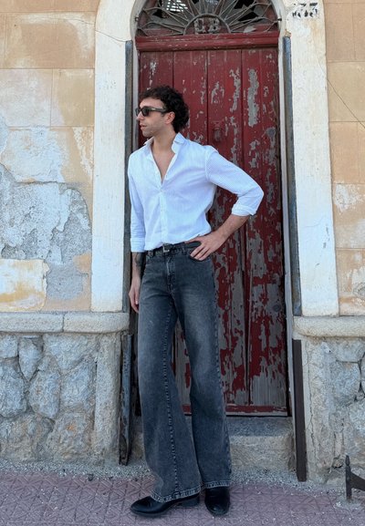 Camisa a rayas blanca y azul, pantalones de cintura alta de pierna ancha negros, zapatos negros; de pie junto a una puerta roja desgastada y una pared de piedra.